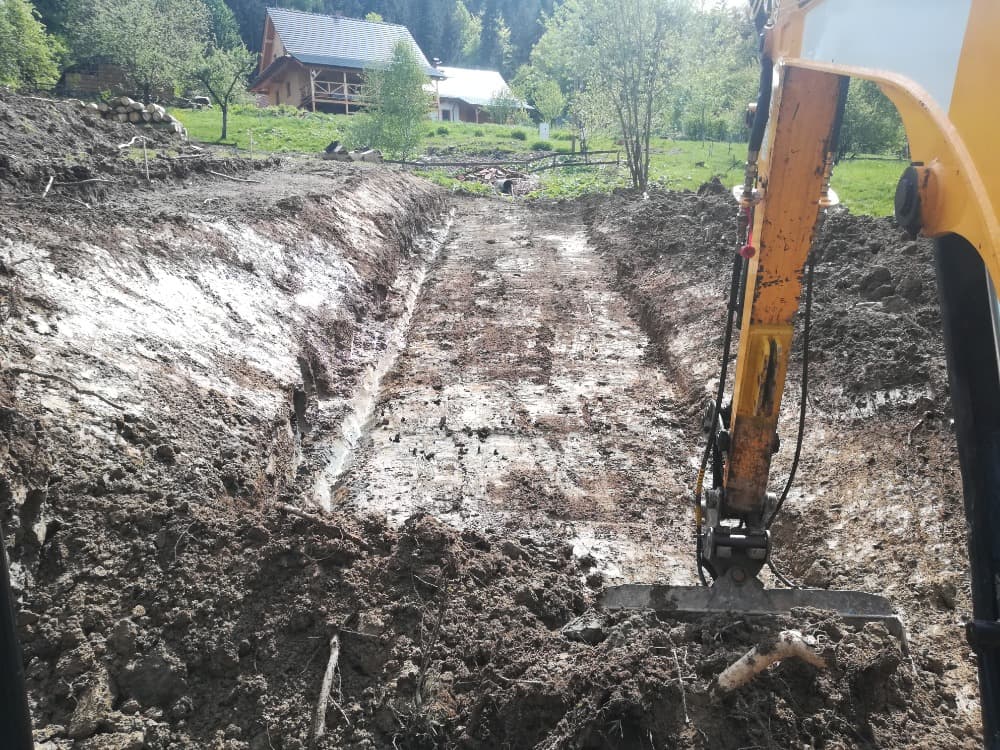 Výkop stavební jámy ve svahu nad chalupou, na kraji výkopu pracuje bagr