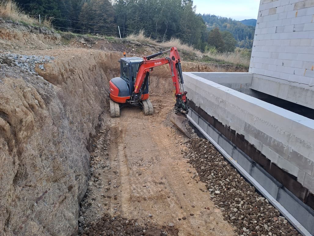 Pásový bagr Kubota v hlubokém výkopu podél základů rozestavěného domu, v pozadí zalesněné beskydské kopce